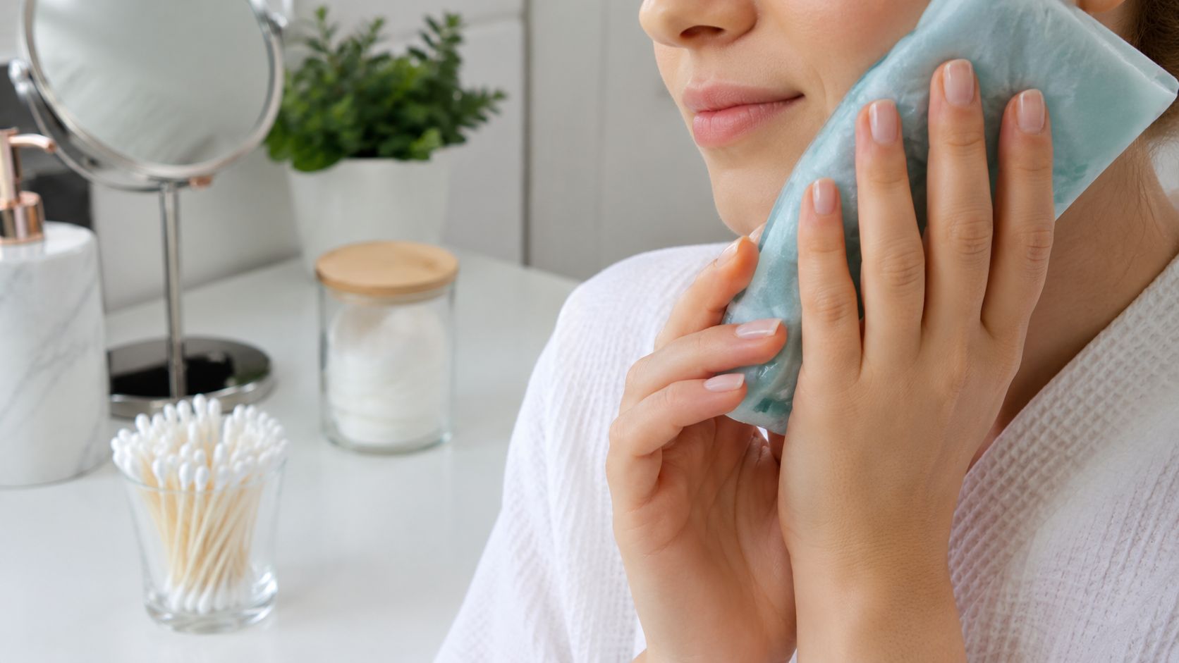 A woman in a bathrobe applying an ice pack to her cheek for tooth pain relief.